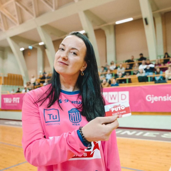 A woman holding a PUTO client card at a basketball game with Rīgas Zeļļi.