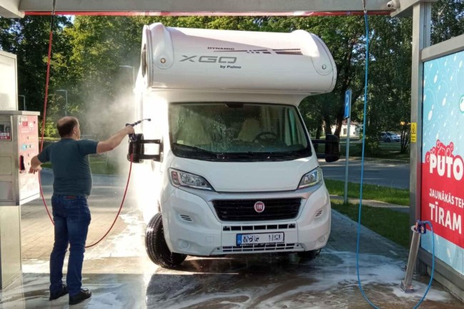 Man washing a white camper van at the PUTO car wash using a high-pressure water spray.